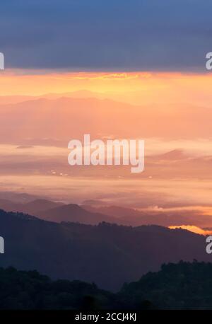 Scenery of idyllic mountain range at sunrise, glowing sunbeam shines through blue clouds on a valley and river in the morning mist. North Thailand. Stock Photo