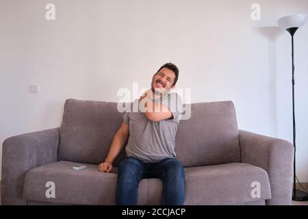 Young man sitting on a sofa suffering from neck pain. Neck and shoulder paint. Stock Photo