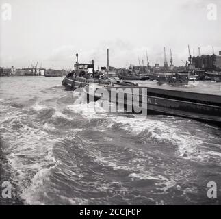 1960s, historical, a steam driven barge pulling an empty steel container vessel up the River Thames, London, England, UK. In this era, the Thames was a working river and warehouses and cranes can be seen lining the riverbank. Stock Photo