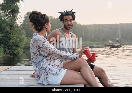 Young couple sitting on a pier with cocktails and talking to each other during summer day Stock Photo