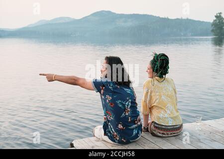 Young man pointing and showing something to his girlfriend while they sitting on a pier and enjoying the view Stock Photo