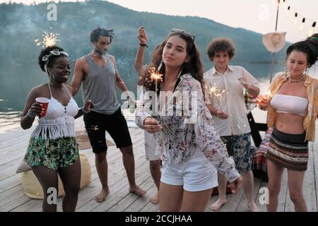 Young woman dancing with sparklers on a pier among her friends at the party outdoors Stock Photo