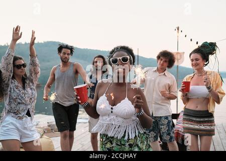 African young woman in sunglasses with glass of beer and sparklers smiling at camera while dancing with her friends at party outdoors Stock Photo