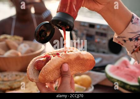 Close-up of woman holding hot dog and adding ketchup on it during dinner Stock Photo
