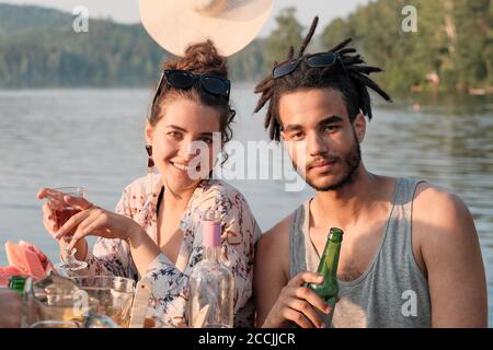 Portrait of young couple drinking beer and smiling at camera while sitting at the table during lunch outdoors Stock Photo