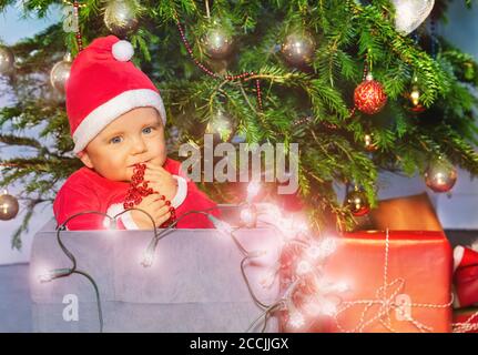 Toddler little boy in Santa Claus hat holding illuminated lamps sitting in box under Christmas tree Stock Photo