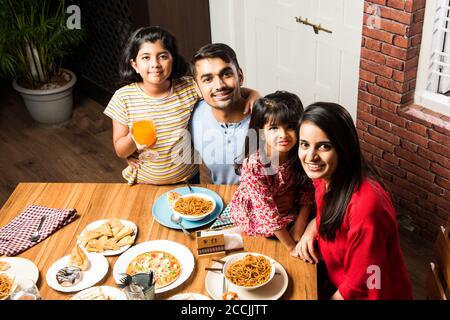 Indian young Family of four eating food at dining table at home or in restaurant. South Asian mother, father and two daughters having meal together Stock Photo
