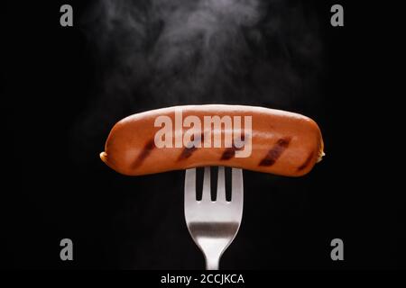 Freshly grilled smoked sausage on a silver fork with rising steam over a black background with copyspace Stock Photo