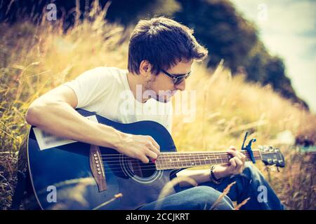 Young man is sitting at a river and playing his western guitar Stock Photo