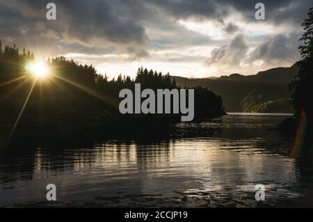 Sunset in Norway over a fjord","Sun setting behind the forest at a fjord. Evening light on beautiful mountians in the background. Midsummer in Scandin Stock Photo