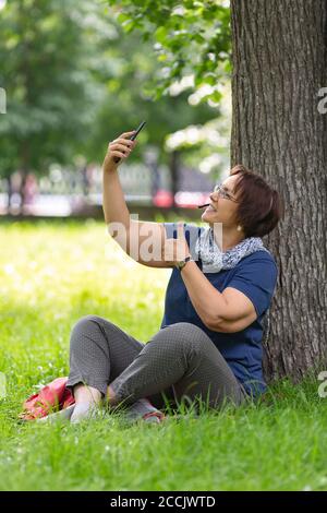 A middle-aged woman using her smartphone, she make a video-call, talks and sends messages. Keeping thump up. Stock Photo
