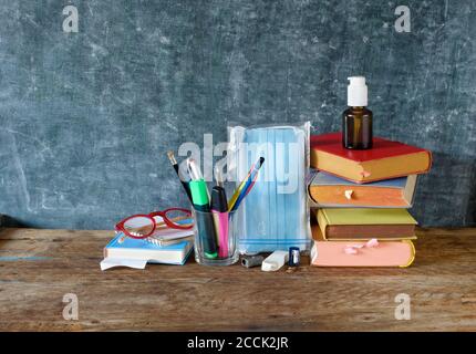 School supplies and COVID 19 prevention items on classroom desk with books,eyeglasses,pens on chalkboard background. Back to school during corona-viru Stock Photo