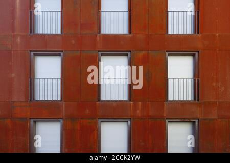 Outdoor exterior front view of modern building with typical rectangular windows, balcony, railing and shutter curtain windows on rust copper facade. Stock Photo