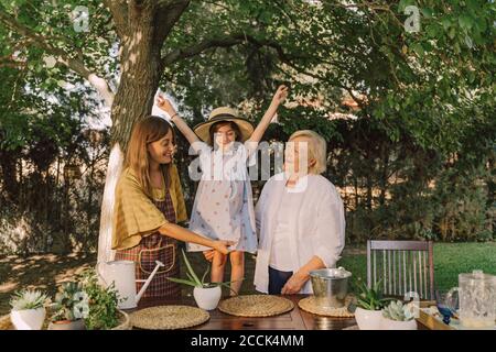 Cheerful girl with arms raised standing with mother and grandmother against tree in yard Stock Photo