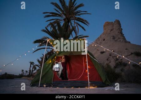 Smiling young tourist woman wearing Hijab looking out of tent at dusk Stock Photo