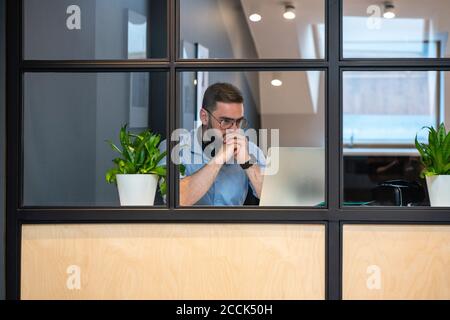Thoughtful handsome male entrepreneur looking at laptop seen through glass wall in illuminated office Stock Photo