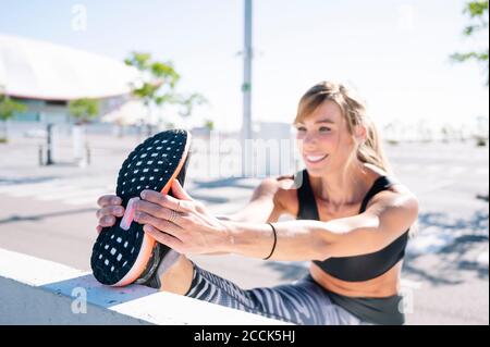 Smiling woman stretching leg on retaining wall in city during sunny day Stock Photo