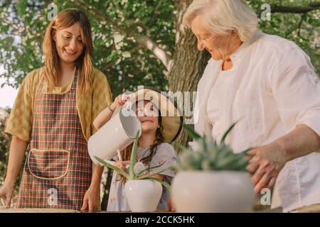 Smiling mother and grandmother looking at girl watering potted plant in yard Stock Photo