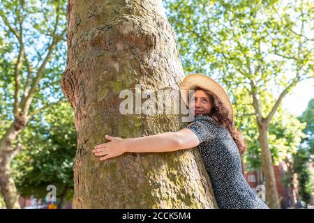 Smiling young woman wearing hat embracing tree in park Stock Photo