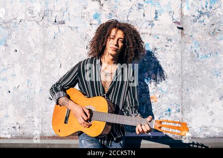 Handsome man with curly hair playing guitar while standing against old wall in city on sunny day Stock Photo