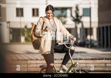 Female student using mobile phone while sitting on bicycle in city during sunny day Stock Photo