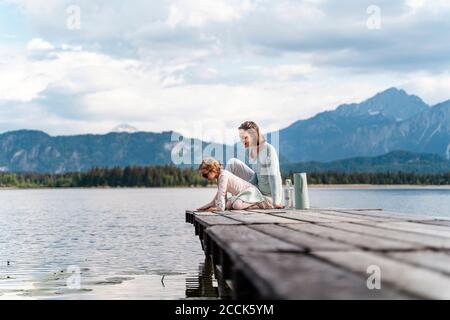 Mother with daughter sitting on jetty over lake against sky Stock Photo