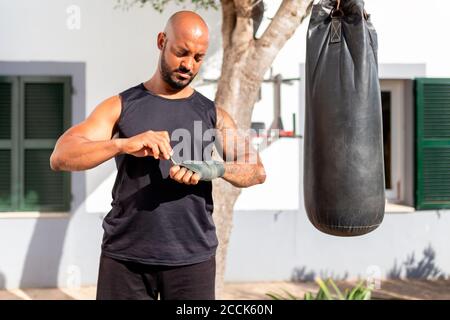 Bald mature man tying bandage on hand while standing by punching bag in yard Stock Photo