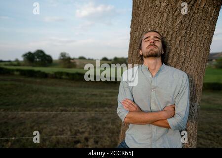 Relaxed young man leaning against a tree trunk in the countryside Stock Photo