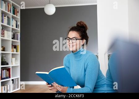 Thoughtful woman looking away while holding book in living room at home Stock Photo