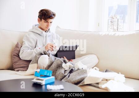 Sick young man using digital tablet while relaxing on sofa in living room Stock Photo