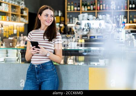 Thoughtful woman holding smart phone while standing at coffee shop Stock Photo