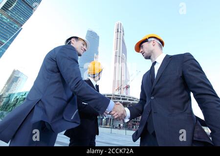 Architects wearing helmets shaking hands at skyscrapers background Stock Photo