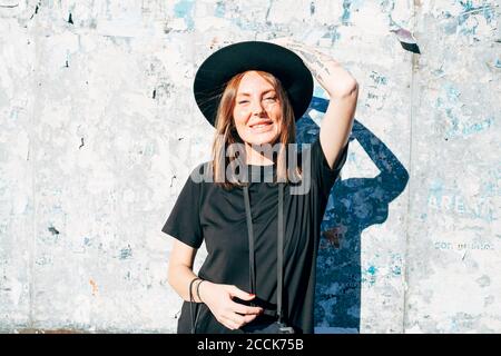 Smiling young woman wearing hat standing against old wall in city on sunny day Stock Photo