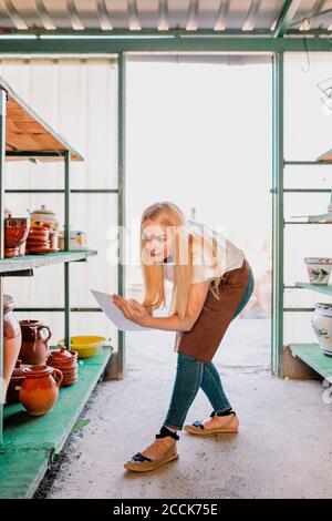 Female owner with blond hair writing on paper while examining potteries in workshop Stock Photo