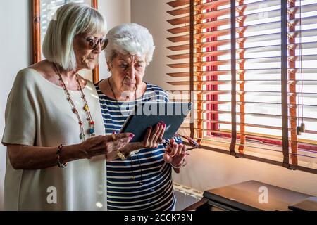 Senior female friends using digital tablet by window at home Stock Photo