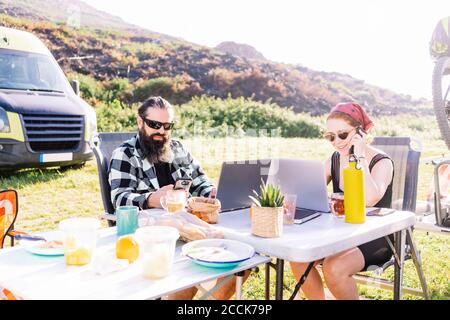 Couple sitting next to camper using laptops on table Stock Photo