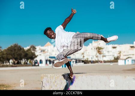 Young man jumping over retaining wall against clear blue sky during sunny day Stock Photo