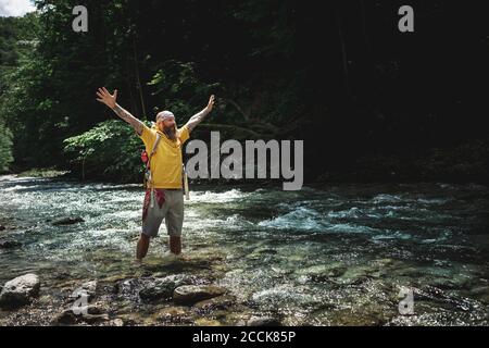 Walker with full beard and yellow hoodie standing in mountain river and raising arms Stock Photo