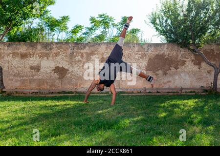 Mature man practicing cart wheel on grassy land against wall in yard Stock Photo
