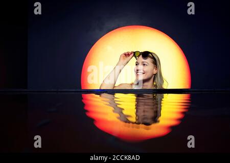 Smiling young woman wearing sunglasses swimming in pool against backdrop of sunset Stock Photo