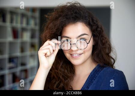 Smiling woman with long brown curly hair wearing eyeglasses at home Stock Photo