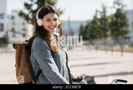 Smiling female student listening music through headphones standing in city on sunny day Stock Photo