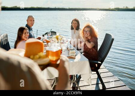 Friends having dinner at a lake with man serving cheese platter Stock Photo
