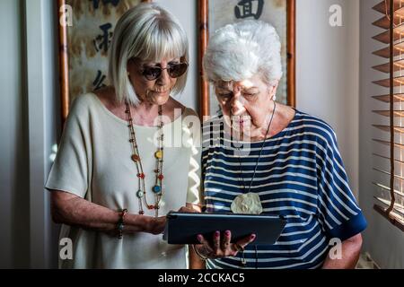 Senior female friends using digital tablet while standing at home Stock Photo