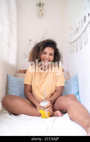 Cheerful young woman with orange juice sitting on bed at home Stock Photo