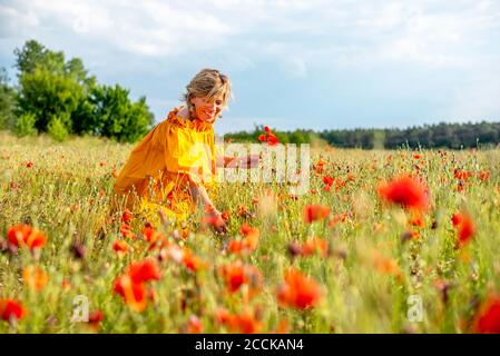 Happy mature woman picking poppies in field against sky Stock Photo