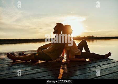 Couple sitting back to back on jetty at a lake at sunset Stock Photo