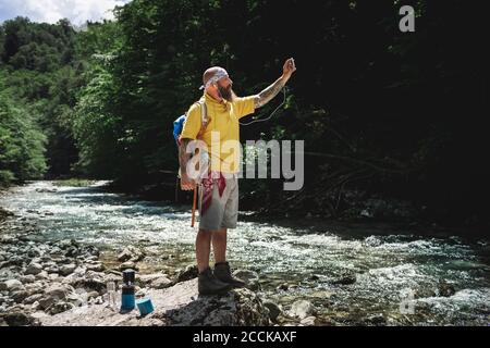 Hiker with full beard and yellow hoodie standing at riverside and using smartphone Stock Photo