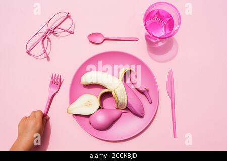 Hand of baby girl picking up fork beside plastic plate with pink-colored pear and banana Stock Photo