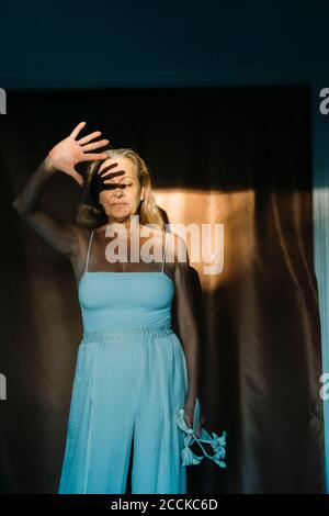 Senior woman shielding face from sunlight with hand while standing against curtain Stock Photo